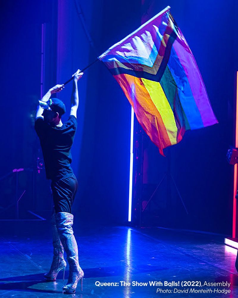 A performer holds a Pride flag aloft on stage. Image from Queenz: The Show With Balls! (2022), Assembly. Photo by David Monteith-Hodge.