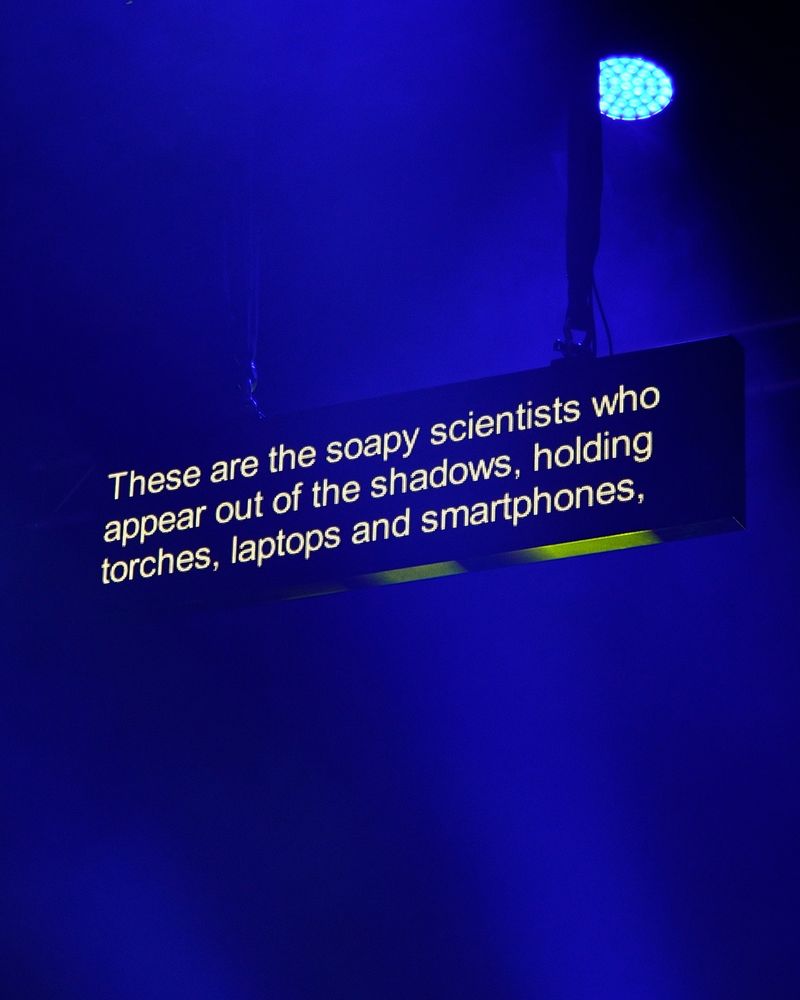 Above a stage, a captioning screen displays the words: 'These are the soapy scientists who appear out of the shadows, holding torches, laptops and smartphones.'