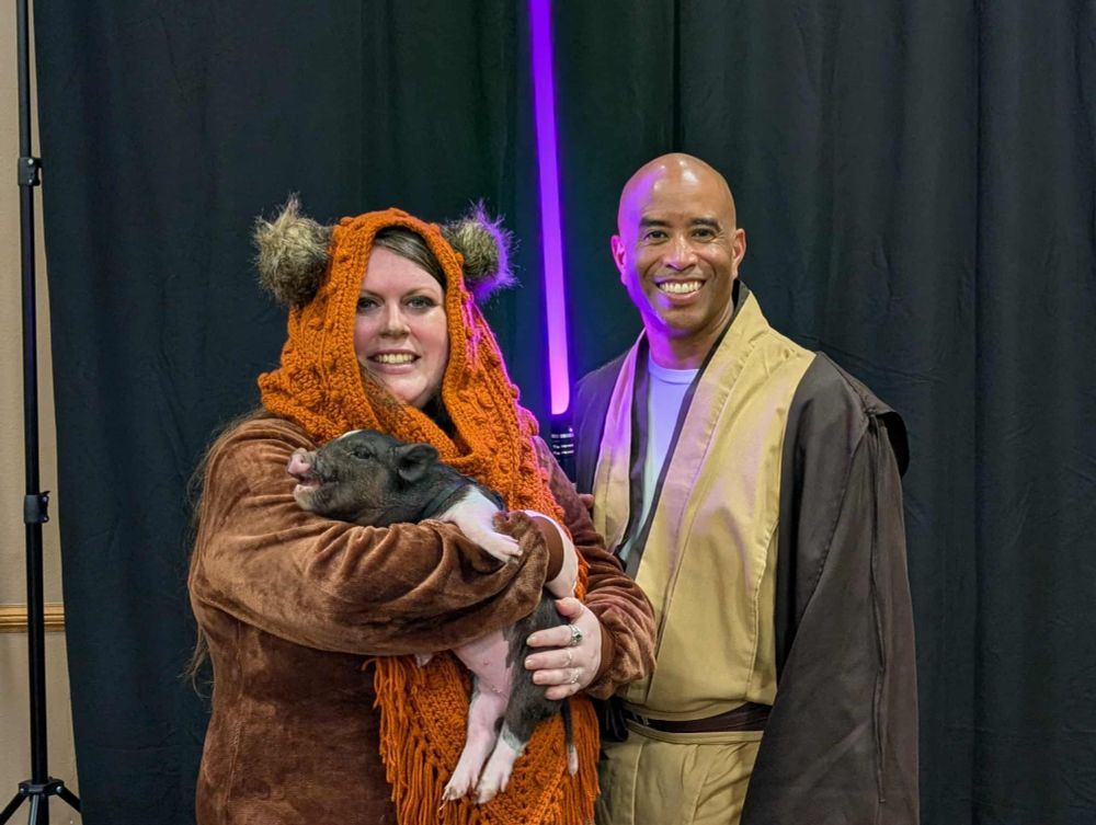 Hannah Solo, a fat cosplayer, in her Wicket the Ewok costume, holding a black and white piglet, next to Mace Windu.