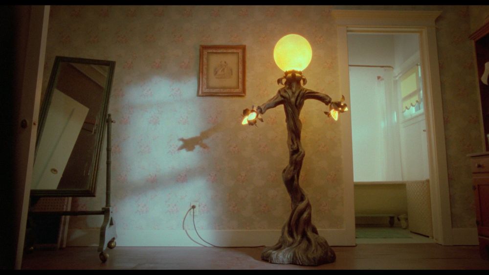 View of a room interior in a typical house. A hideous floor lamp is prominent. It looks like a skinny tree twisted into the rough shape of a person with arms outstretched. A large spherical bulb cover is lit where the "head" might be, while two smaller bulbs are present in each of the "hands".