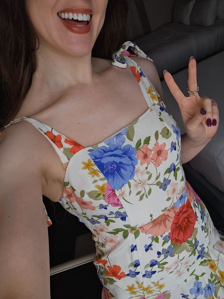 Sam is wearing a white dress with flowers of all different colors on it. She's smiling and giving the peace sign.