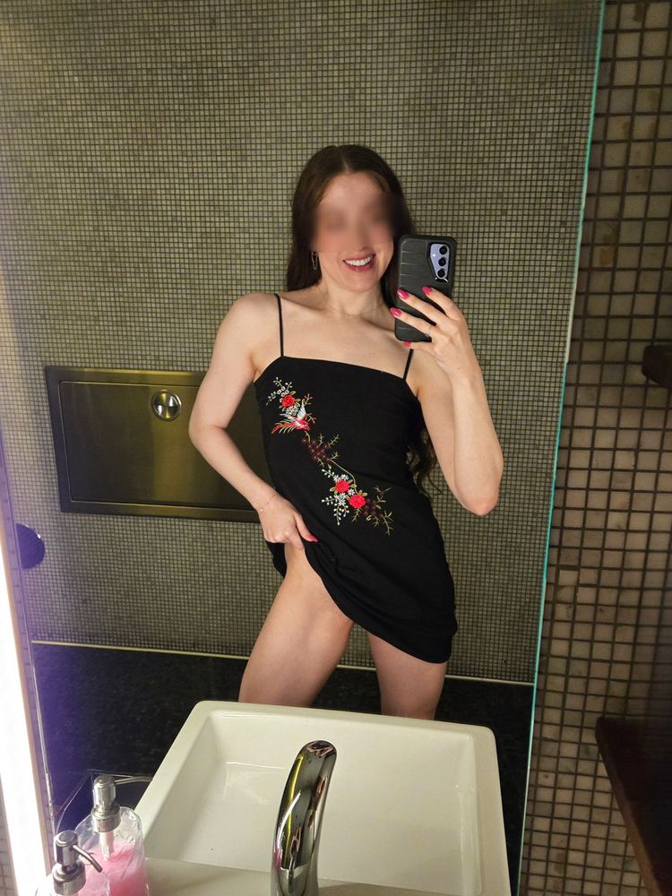 Sam is wearing a short black dress with some red flowers on it, smiling, looking into a mirror and lifting her dress to show one leg. 