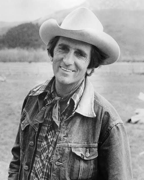 Black and white photo of Harry Dean Stanton ,wearing a cowboy hat and jean jacket and smiling. 