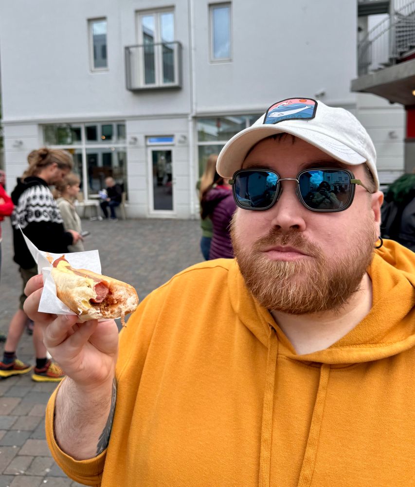 Bearded person holding up a lamb hotdog with their mouth full of food. The Traditional Icelandic hot dog with Icelandic ketchup, Icelandic mustard, remoulade sauce, deep fried onions and raw onions.