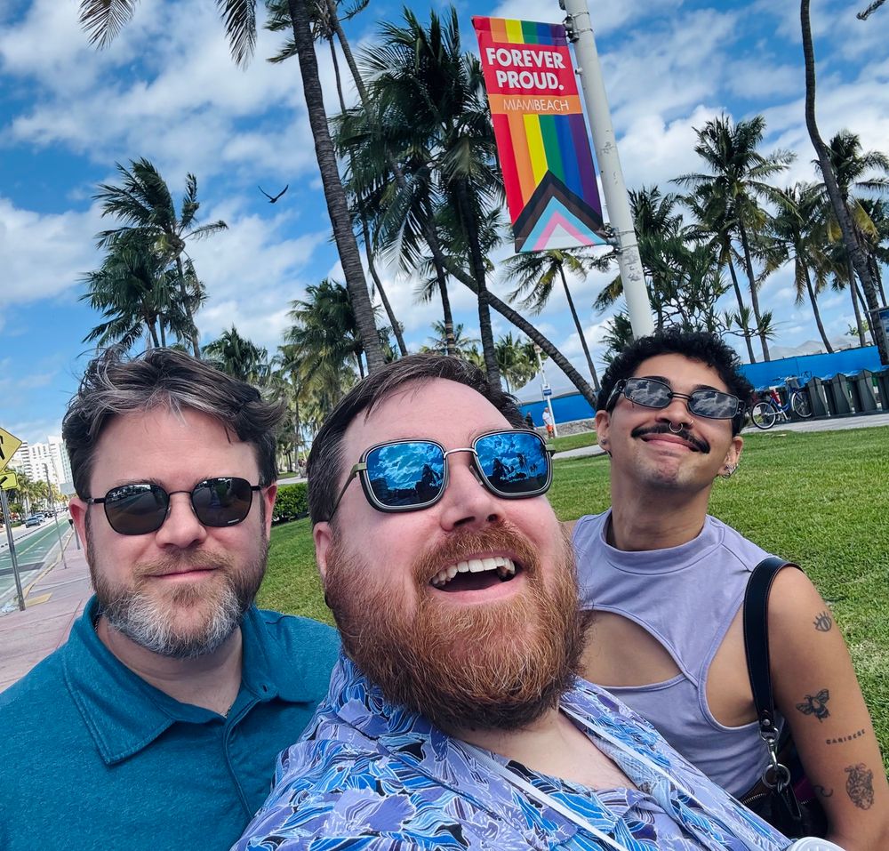 3 sunglassed hotties smiling for the camera in front of a rainbow flag in Miami Beach. 