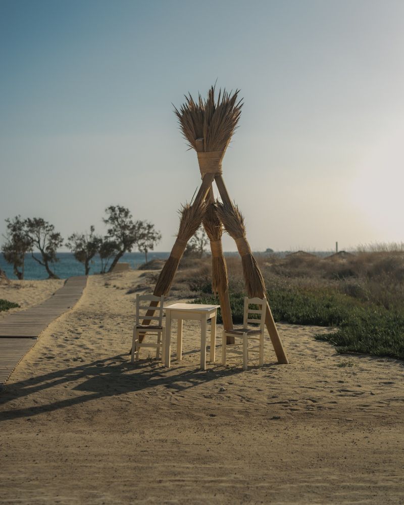 A rustic beach scene with a teepee-like structure made of dried reeds, standing on sandy ground. The structure is triangular, bound together at its peak, with a small white table and chairs placed nearby. In the background, a wooden boardwalk leads towards the ocean, with sparse trees and vegetation visible. The sky is a soft blue-green, creating a serene coastal atmosphere.