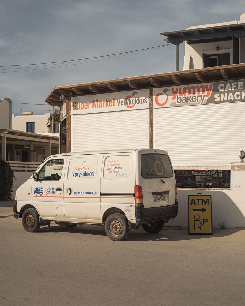 A white delivery van for 'Super Market Verykokkos' parked in front of a storefront with roller shutters. The building has signage for 'Yummy Bakery' and 'Cafe Snack'. An ATM sign is visible nearby, and the scene appears to be in a sunny, urban or suburban setting with buildings and a cloudy sky in the background. The van has contact numbers and a website printed on its side. 