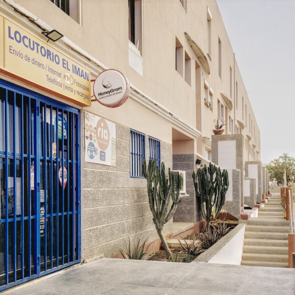 A beige building with blue-barred windows and doors houses a business called "Locutorio El Iman." The storefront sign advertises services such as money transfers, internet, photocopies, and phone sales and recharges. There are also signs for MoneyGram and Ria Money Transfer. In front of the building, there are several tall cacti and a walkway with steps leading up to other entrances into a Fuerteventura project complex.
