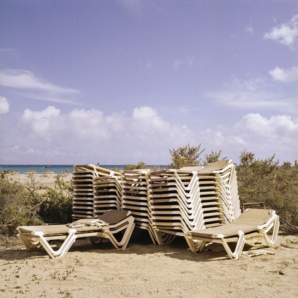 Two large stacks of white plastic sun loungers with brown cushions sit on a sandy beach. Behind them, more sun loungers are visible further down the beach towards the ocean, which is under a partly cloudy sky. Dry, sparse vegetation grows around the stacks.
