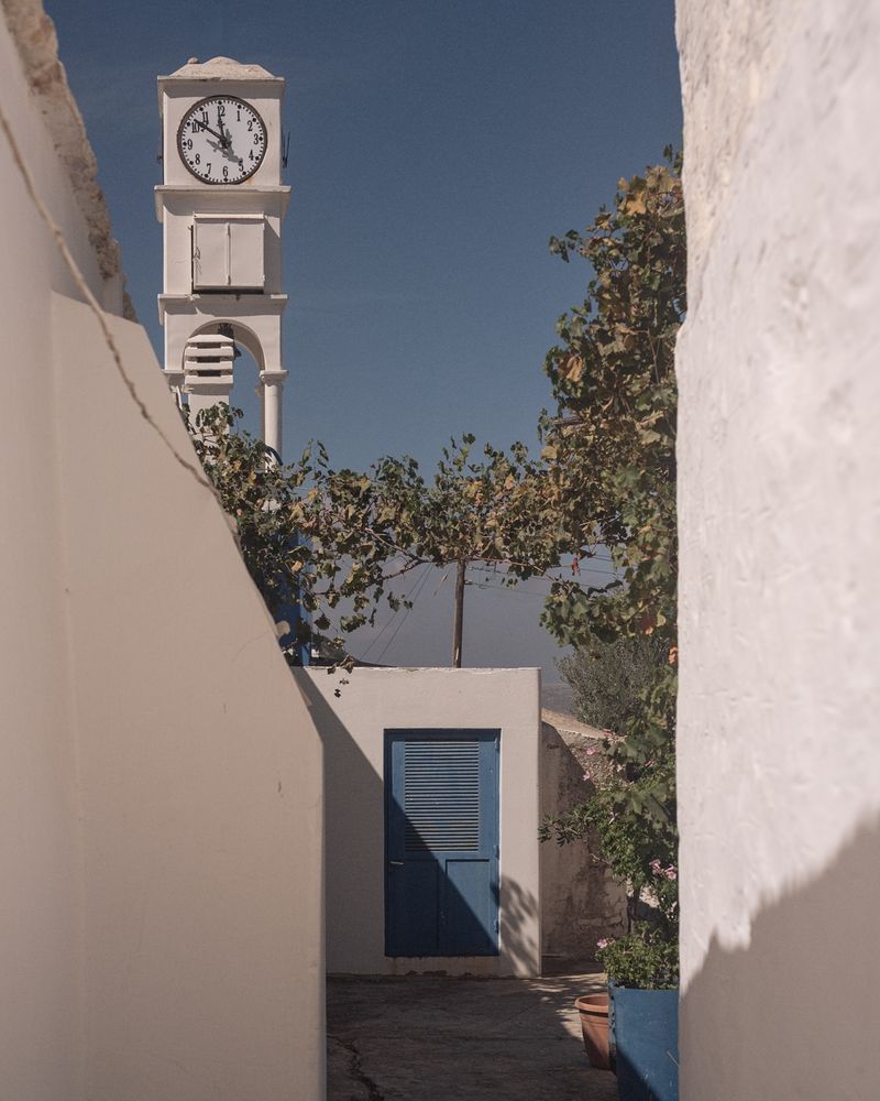 A white stone architectural scene featuring a tall clock tower with a white analog clock showing the time near 10:50. The clock tower is framed by white walls and surrounded by green leafy trees. In the foreground, a blue louvered door is visible, set against white walls with soft shadows. The scene has a Mediterranean or Greek island aesthetic, with clean white surfaces and a deep blue sky in the background.