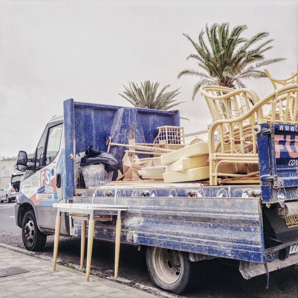 A blue flatbed truck parked on a street, loaded with various discarded furniture pieces including chairs, cushions, and a small table, with palm trees and a cloudy Fuerteventura Sky in the background.