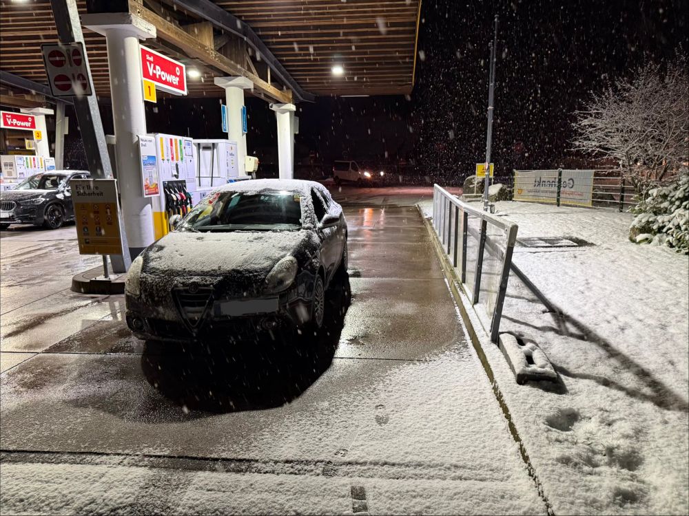 Ein verschneiter schwarzer Alfa an einer Tankstelle. Es ist dunkel und schneit heftig. 