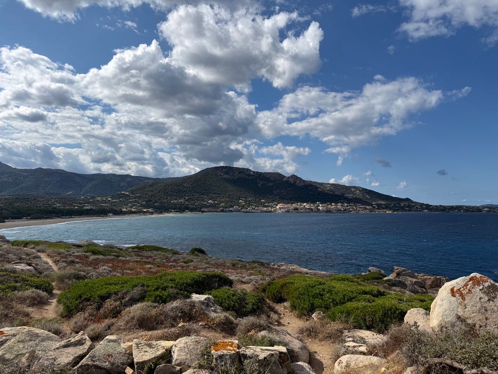 Blick über eine Meeresbucht (Korsika) mit steiniger Landschaft im Vordergrund. Die Sonne scheint, es ist aber auch bewölkt. 