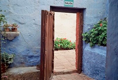 Bright blue walls made from volcanic stone inside the Santa Catalina Monastery are lined with green plants. A heavy, open, wooden door looks out into a sunny passageway onto blooming plants. The sign above the door reads “salida.” 