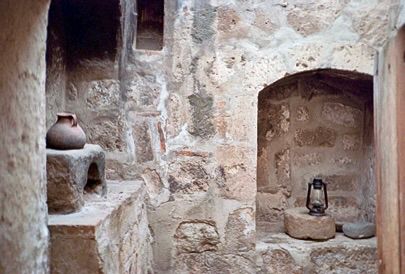 This image shows a rudimentary kitchen inside one of the cells of the Santa Catalina Monastery in Arequipa, Peru. There is an oven made from volcanic rock with a pottery pitcher sitting on it. Across from that there is an alcove with a couple of grinding stones, one of which has a lantern on top of it. 
