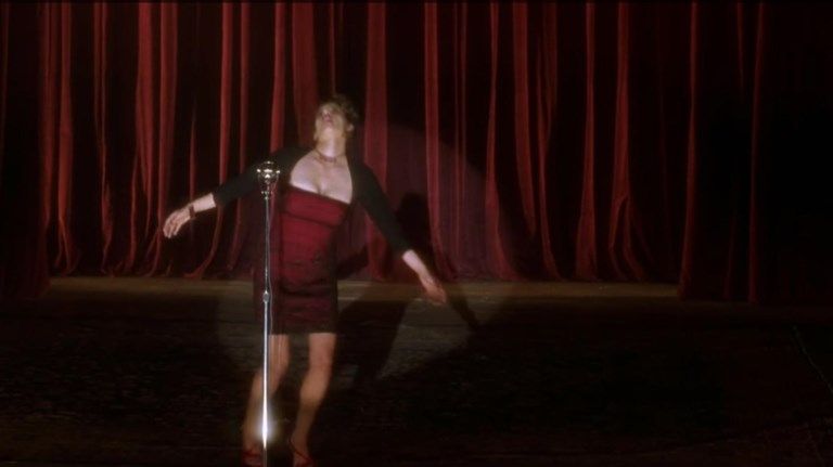 In a still from Mulholland Dr., a woman in a dark red dress at an upright microphone begins to fall. The spotlight shines on her. A dark red curtain fills the stage's background.