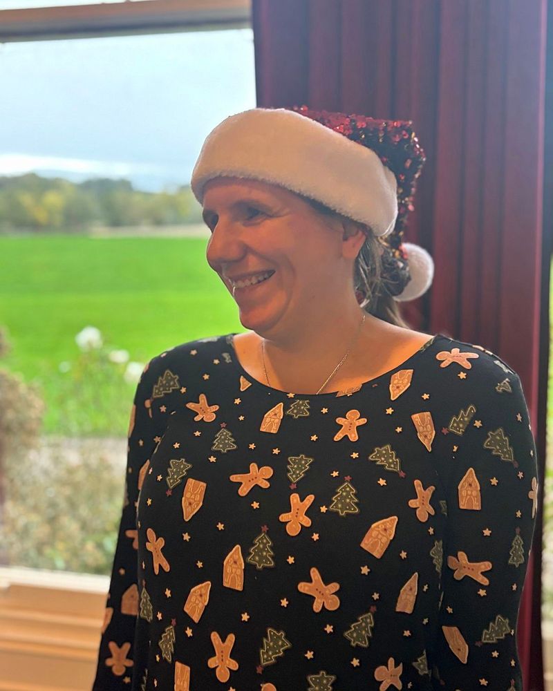 A young woman smiling at the camera wearing a Santa hat and a festive top decorated with Christmas trees and gingerbread men.