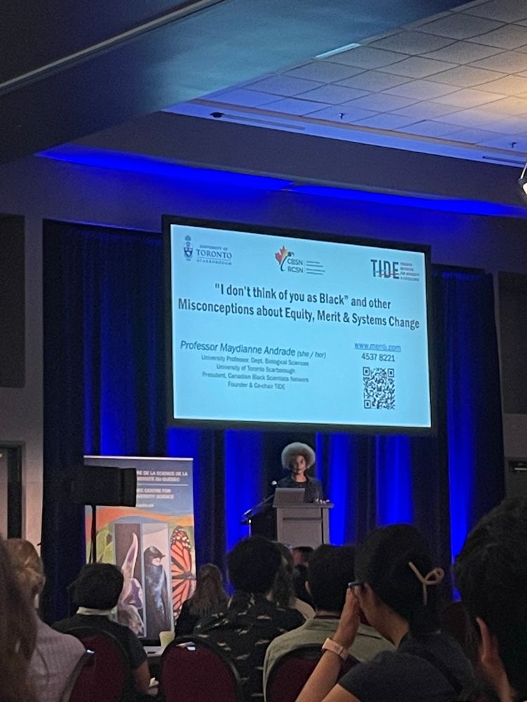 Maydianne is standing on stage behind a podium. The project screen shows a slide that reads the title of the talk “I don’t think of you as Black: Misconceptions about equity, merit and systems change”