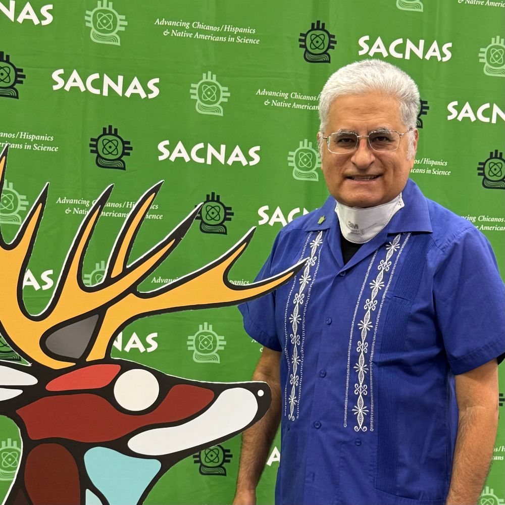 Alberto Roca in blue guayabera shirt & glasses in front of SACNAS green banner. Sign alternates between “SACNAS” in white font and New Mexico glyph logo in black. At Alberto’s side is cardboard cutout statue of 2025 themed antlered deer colored in white, brown, and light blue pattern.