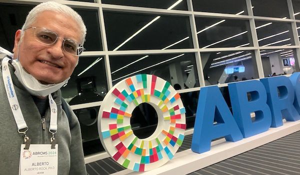 Selfie photo by large sign that spells out ABRCMS in blue letters. Sign also has their logo which is circle of white background interspersed with red, green, blue, orange wedges. Roca is smiling at camera; and, he has gray hair, wearing gray sweater, and conference name badge.