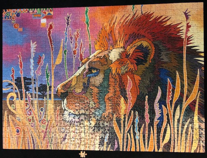 Photo of 1000 piece puzzle depicting an illustration of the side profile of lion lying in tall grass. The artist's painting style has many geometric shapes filling in the yellow and orange grass blades. The lion's fur coloring is interpreted as jagged patches that range in color from grey-brown to tan in the animal's face while the mane is colored dark orange to red to dark green. The background is a blue & purple tinted savannah evening sky where the artist has triangle, square, and circles in the top left corner in various colors such as green red that match the colored tips of the foreground grass that is reaching to the sky above the lion's head.