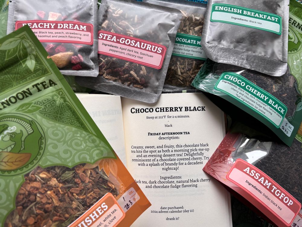 Bags of different blends of loose leaf black tea from Friday Afternoon Tea are piled around and on a book open to an entry giving ingredients and brewing suggestions for one of the teas in the pile