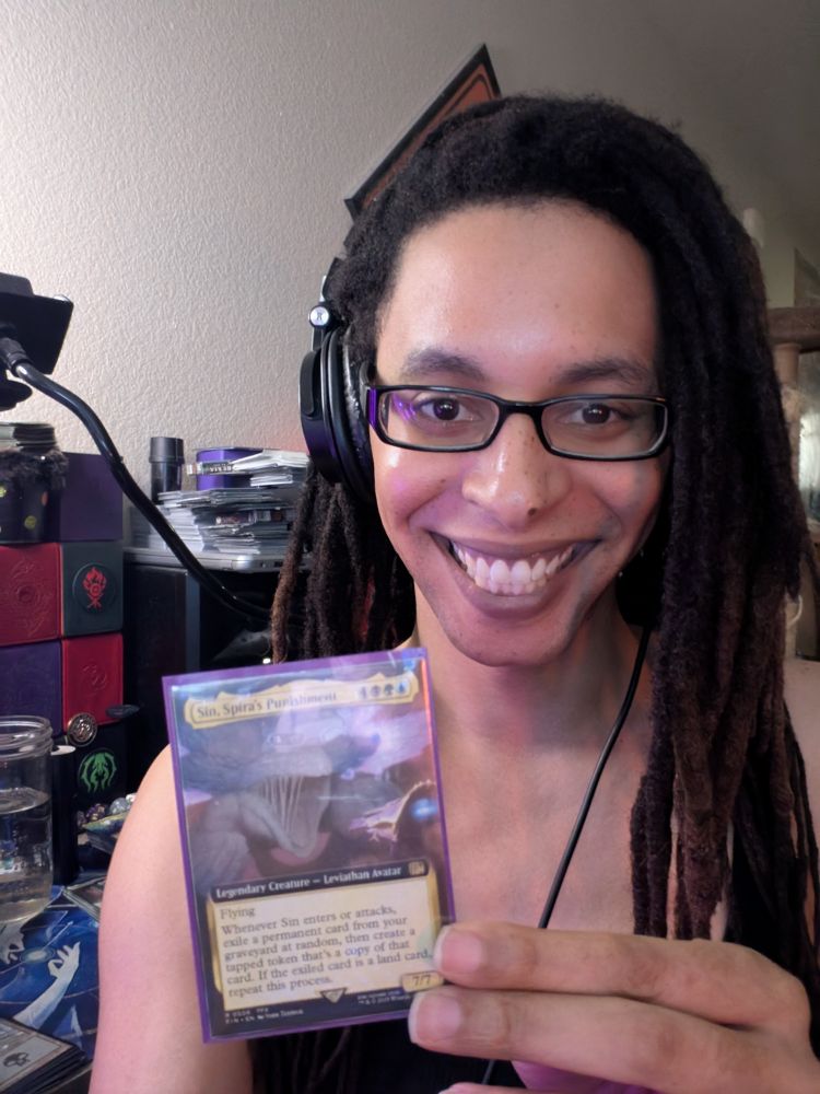 A selfie angle photo of Empress Quinn in front of faer desk holding a copy of the magic the gathering card Sin, Spira's Punishment. We are prepared to game. 