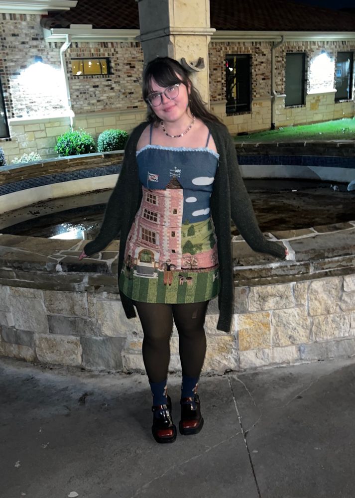 Clare dressed in a short dress, cardigan, and tights posing outside 