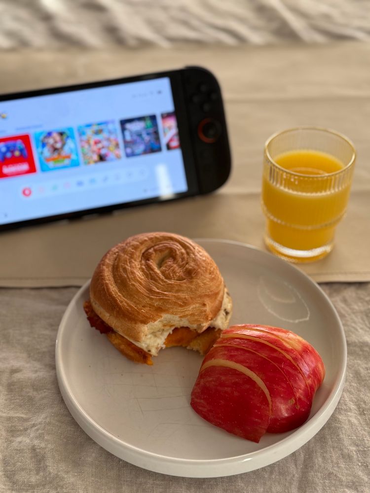 Apple slices on a plate with a breakfast, croissant sandwich, orange juice, and Nintendo switch on a table