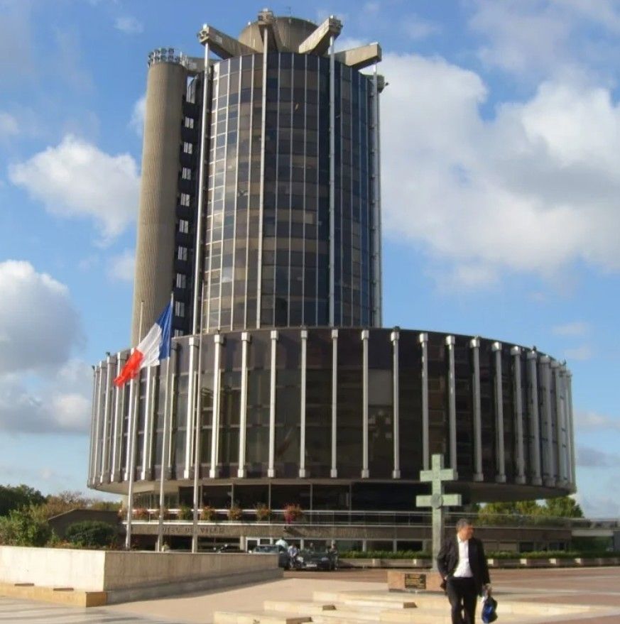 1970s modern building formed by a low wide cylinder and a much taller narrower one. There's a French flag and a cross of Lorraine in the foreground and a middle aged man.