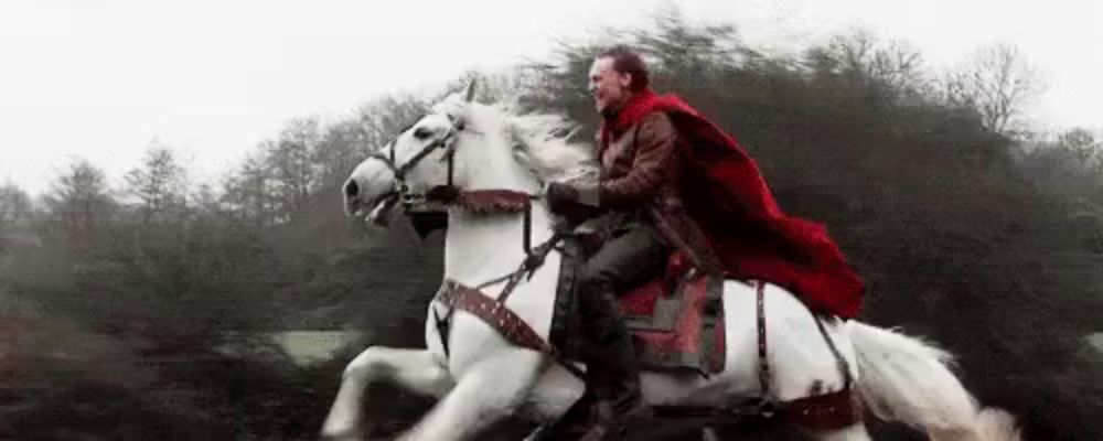 a man in a red cape is riding a white horse through a forest .