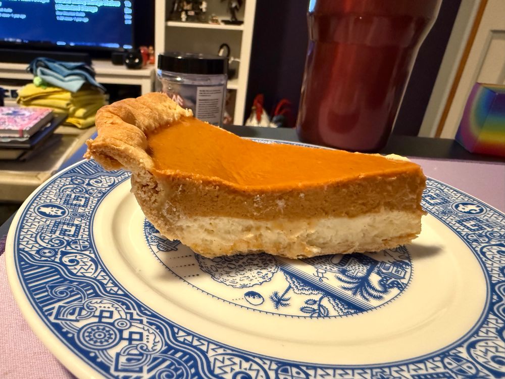 A slice of pumpkin cheesecake pie on a plate. It has a vanilla cheesecake layer on the bottom and pumpkin custard on top. 