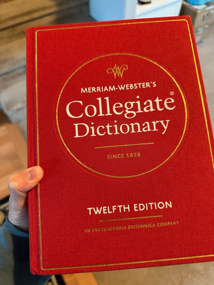 A white hand holds up the twelfth edition of Merriam-Webster’s Collegiate Dictionary. It is bound in red cloth with gold foil. 