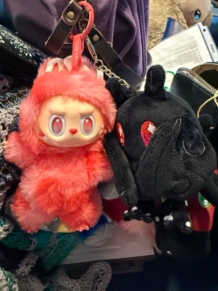 A hot pink “Love” Labubu (from the Big Into Energy) line hangs next to a micro Squishable Mothman, off the zipper of a purple project bag. The whole area is a riot of knitting related mess that we won’t talk about right now, but that I desperately need to organize. 