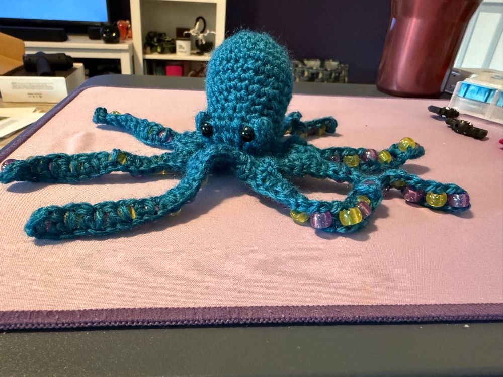 A small blue crocheted octopus sits on a lavender desk mat. He has yellow and pink translucent beads in his arms; to act as things to forget with. 