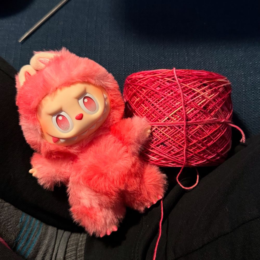 A hot pink labubu lays next to a ball of yarn in a strikingly similar color. 