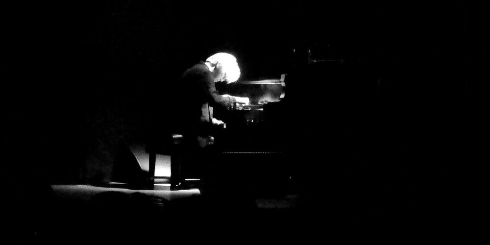 Das Foto ist schwarz und weiß gehalten; es ist größtenteils dunkel. Etwas bildmittig ist Licht zu erkennen. Im Lichtschein sitzt ein Mann an einem Klavier, sein Haar ist weiß.