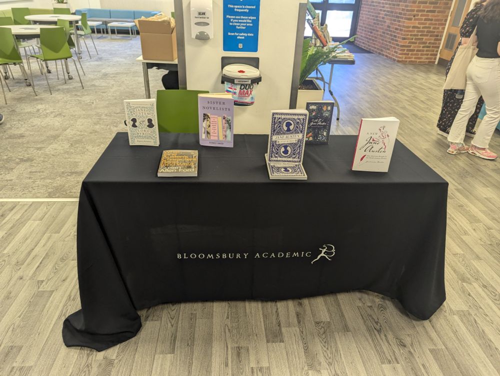 Photograph of a table with a Bloomsbury tablecloth on it, along with six books all on Jane Austen 