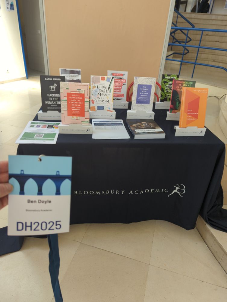 Photograph shows a Bloomsbury Academic book stand with ten digital humanities books on it, along with some flyers. There is a hand holding a DH 2025 lanyard in the foreground.