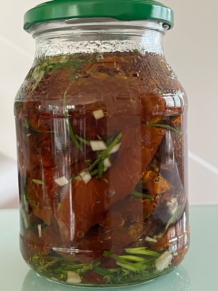 Das Foto zeigt ein Glas mit eingelegten getrockneten Tomaten in Öl mit Knoblaucheürfeln, Thymian und Rosmarin.
Das Glas füllt das Bild fast ganz aus und steht vor hellem Hintergrund auf einer grünlich schimmernden Glasplatte.