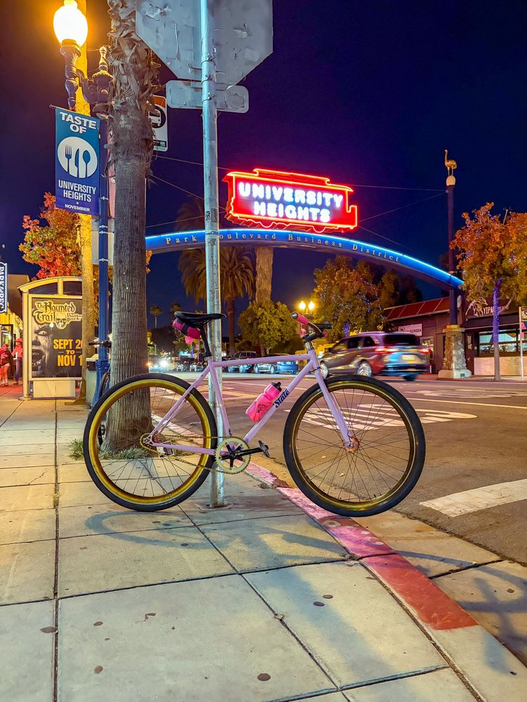 my new lavender state 4130 fixed gear frame with all the parts i collected to build a tracklocross bike installed sitting below a neon sign that says university heights 
