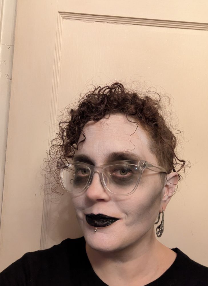 Woman in 3/4 profile with chicken foot earrings and clear glasses, wearing sheer white makeup, black eye sockets, grey contour under cheekbones, and black lipstick.