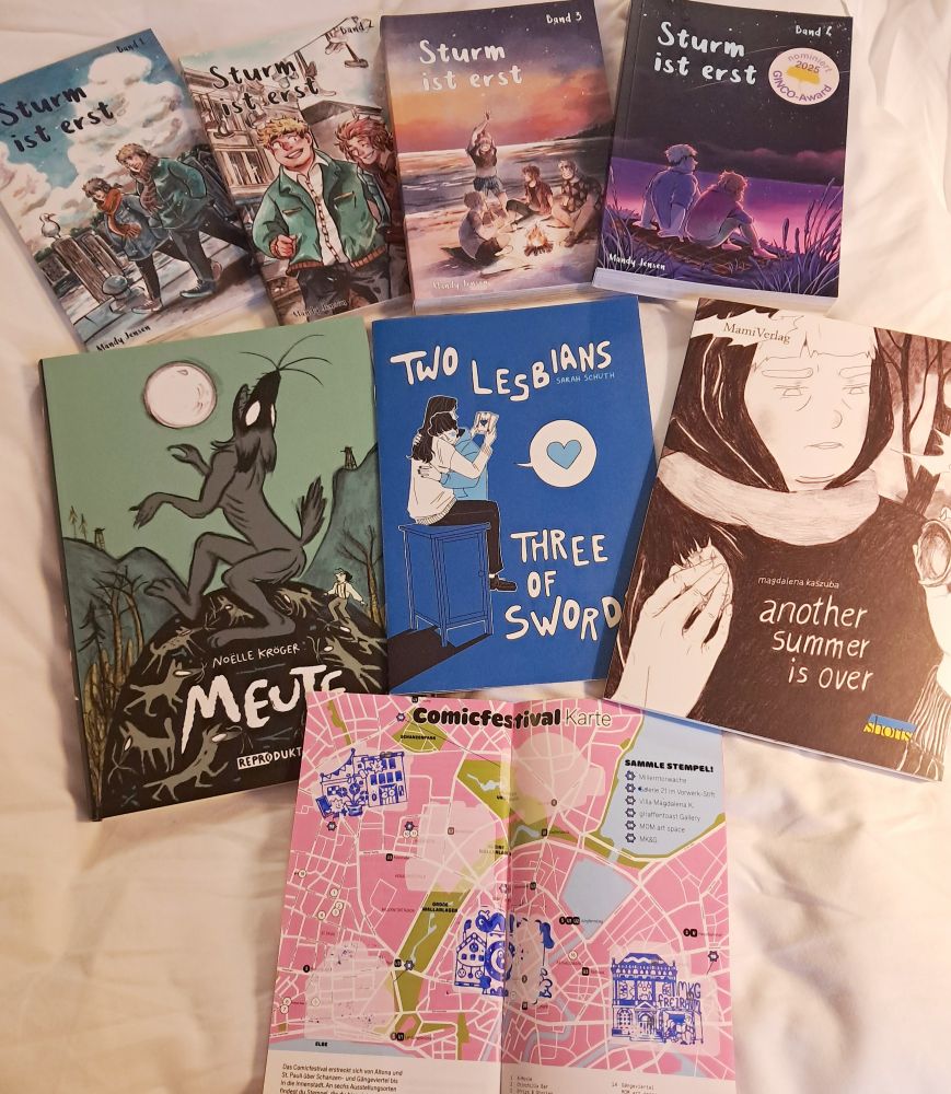 Ein Haufen Comics ausgebreitet auf einer Bettdecke. Die ersten vier Bände Sturm ist erst von Mandy Jensen, Meute von Noëlle Kröger, Two Lesbians Three of Swords von Sarah Schuth, Another Summer is Over von Magdalena Kaszuba und das Programmheft des Comicfestivals, aufgeschlagen auf der Seite mit der Karte, drei von fünf Stempeln sind gesammelt.