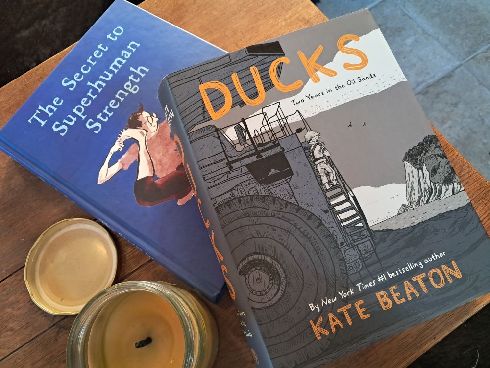 Two books on a wooden tabletop next to a beeswax candle in a glass jar. It's The Secret to Superhuman Strength by Alison Bechdel and Ducks by Kate Beaton.