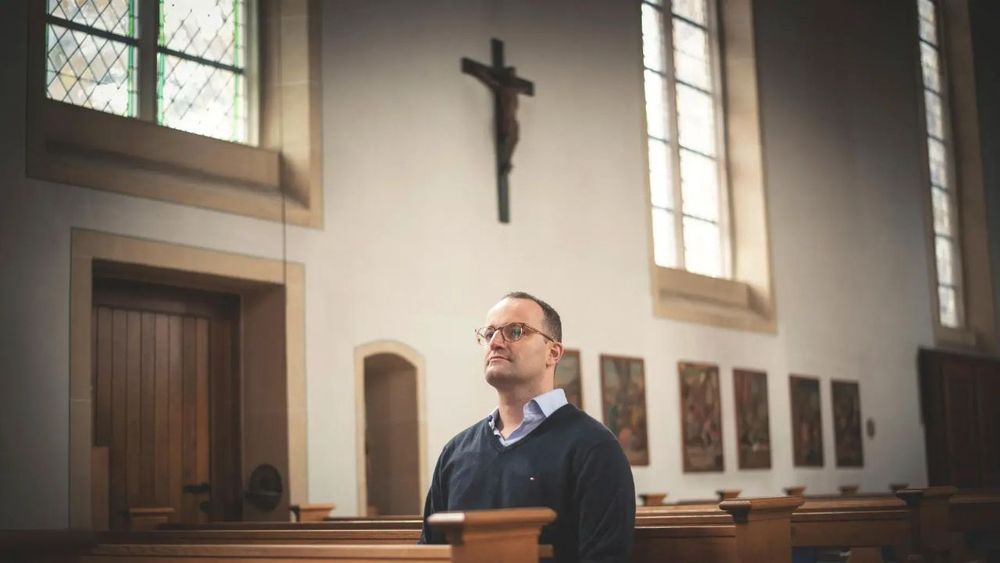 Man sieht das Innere einer katholischen Kirche. Im Zentrum des Bildes Jens Spahn, der entweder kniet oder sitzt und demütig reinblickt. Über seinem Kopf das Kreuz mit dem gehängten Jesus. Im Bild überwiegen braun-weiß Töne.