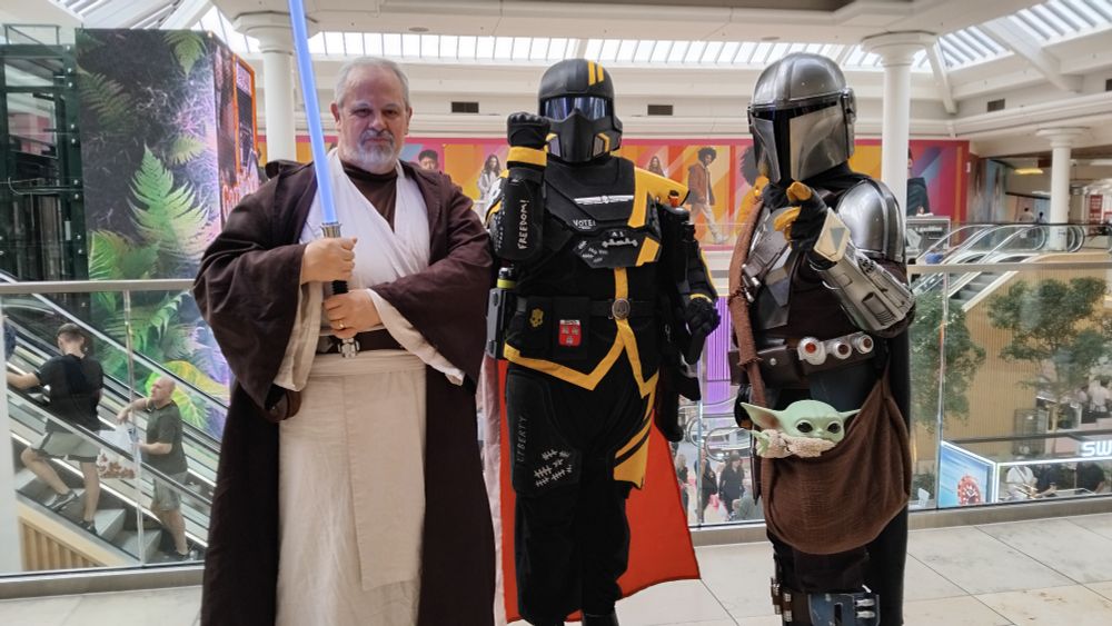 A Helldiver standing in between two cosplayers: Ben Kenobi, a Jedi wearing brown and white robes whilst holding a blue Lightsaber with both hands. And Din Djarin, a Mandalorian clad in silvery armour and a brown body suit. A small green alien, Grogu, is hanging out of his satchel.