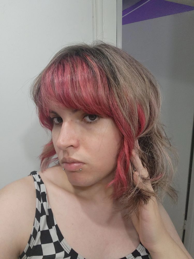 alice taking a selfie. she has dark red/pink bangs with that color mixed into her blonde hair.