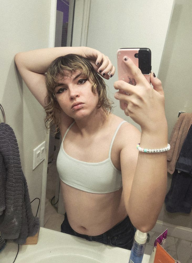 alice taking a photo of themself in the mirror, she is wearing a sports bra and jeans.