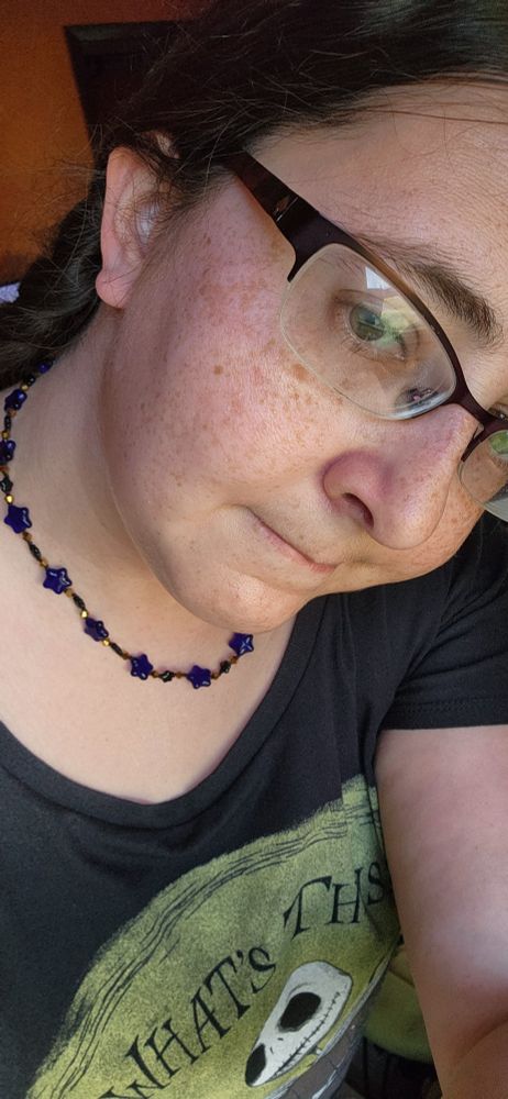A white woman with glasses and braids and a Jack Skellington t-shirt is looking at the camera to show off her necklace. It's dark blue  glass stars with smaller black stars and gold beads.