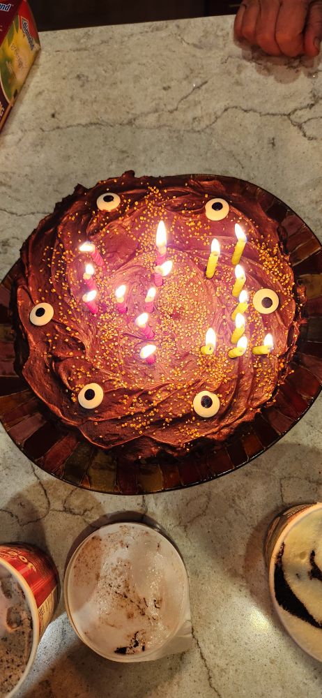 A cake with two ice cream pint containers on a counter top. The cake has chocolate frosting, gold sprinkles, and sugar eyeball decorations. There are lit red and yellow candles forming the number 41.